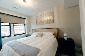 Een slaapkamer met een groot bed, een raam met jaloezieën, een wanddecoratie en een nachtkastje met een lamp.