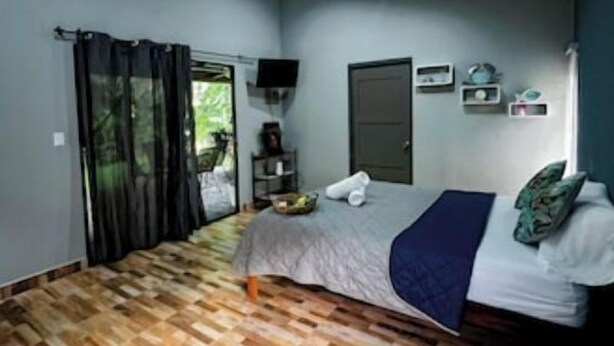 Un dormitorio con piso de parquet de madera, una cama con manta azul, un televisor en la pared y una puerta con un pequeño estante encima.