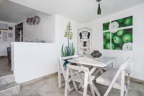 A dining area with a white table and chairs, a cactus, a clock, and a wall with green apple paintings.
