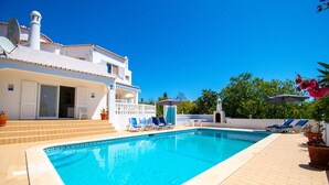 A rectangular swimming pool with a tiled edge, surrounded by a brick patio. There are lounge chairs and umbrellas on the right side. - Casa Pazovida - Walk to Carvoeiro, a perfect blend of convenience, comfort and luxury! (Carvoeiro)