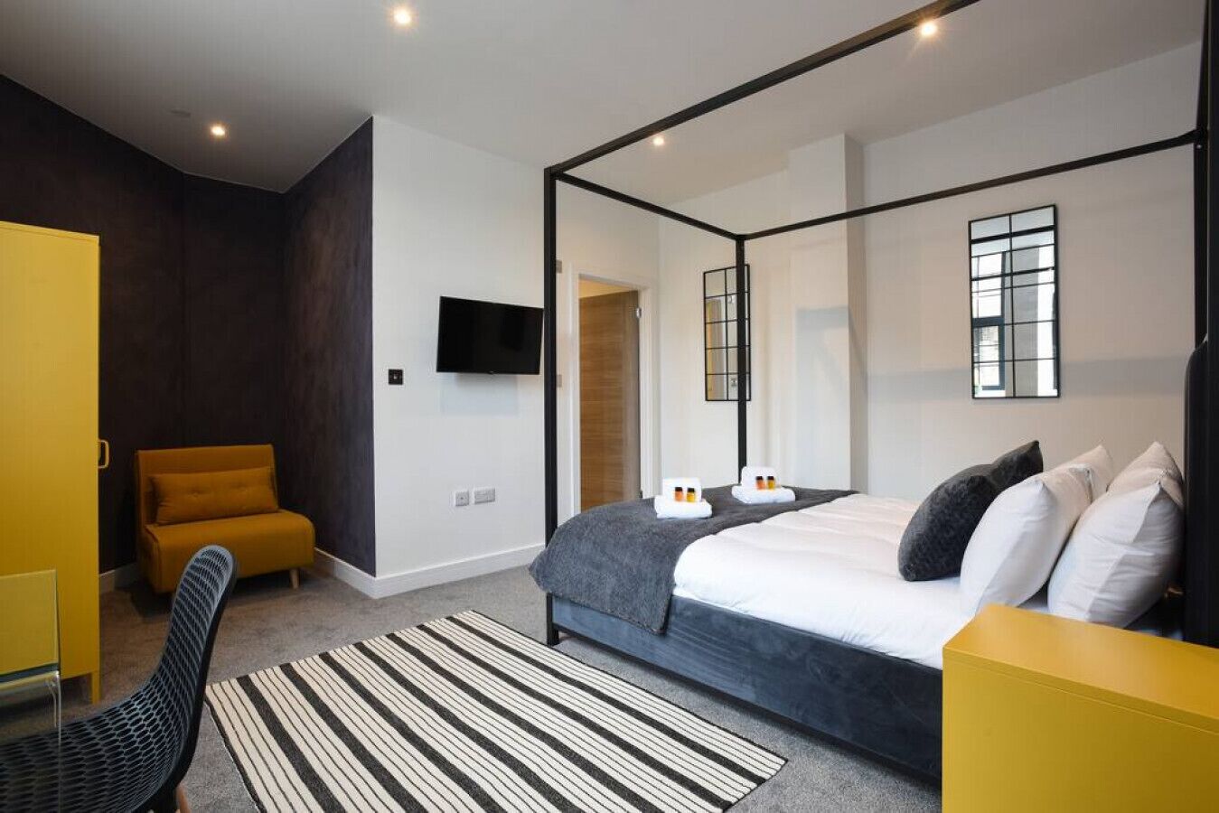 Een moderne slaapkamer met een hemelbed, een televisie aan de muur en een gele fauteuil.