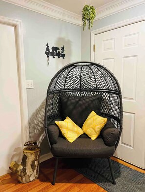 Een zwarte wireframe stoel met gele kussens, een klein houten krukje en een hangplant in een kamer met witte muren en een deur.