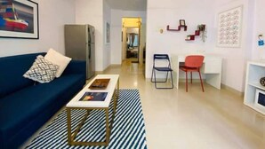 A modern living room with a blue sofa, a coffee table, and a striped rug. There is a desk with two chairs and a ceiling fan. - Nature Inspired Sun Drenched 1BR in Bandra (Mumbai)