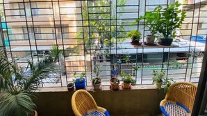 A balcony with two wicker chairs, potted plants, and a view of the street and buildings. - Nature Inspired Sun Drenched 1BR in Bandra (Mumbai)