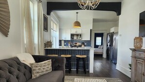 A modern living room with a dark grey sofa, a patterned throw pillow, and a woven rug. A chandelier hangs from the ceiling, and a kitchen with dark blue tiles and white cabinets is visible in the background.