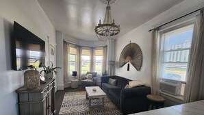A living room with a sofa, coffee table, and decorative items. There is a chandelier, a large mirror, and a window with blinds.