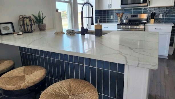 A modern kitchen with a marble countertop, a stainless steel stove, and a microwave. There are two wicker stools and a potted plant on the counter.