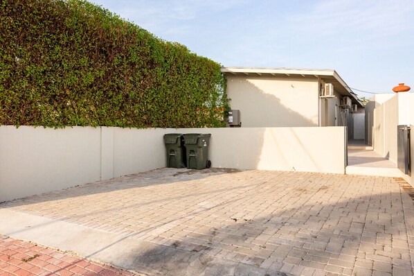A paved courtyard with a white wall and a hedge. There are two green bins and an air conditioning unit on the wall.