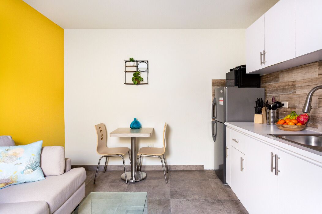 Una cocina compacta con armarios blancos, un refrigerador gris y un lavamanos. Hay un área de comedor con una mesa y dos sillas.