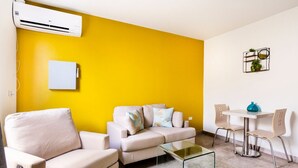 A living room with a yellow wall, a beige sofa, a glass coffee table, and a small dining area with a white table and two chairs.