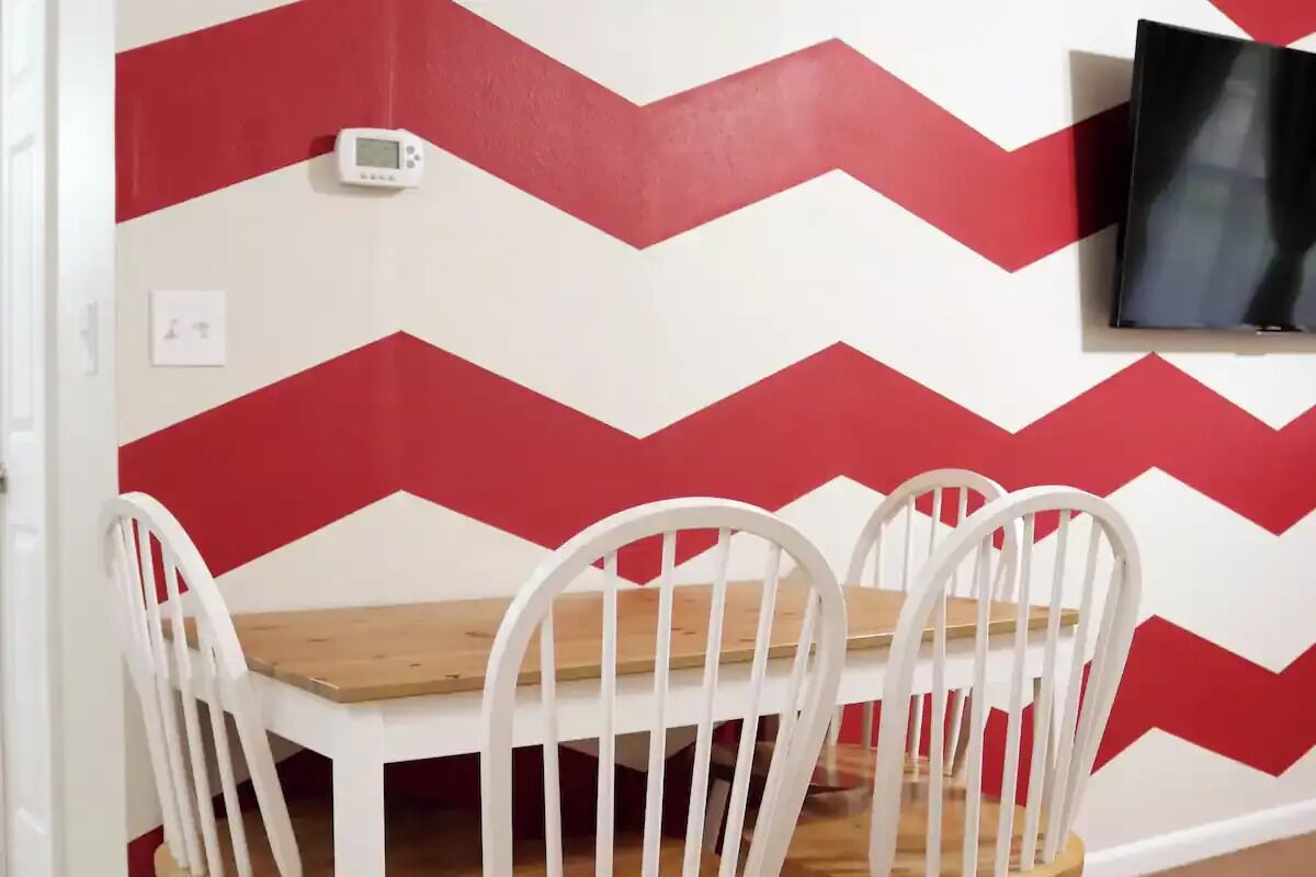 Un comedor con mesa de madera y sillas blancas, pared con estampado blanco y rojo en forma de tablero de ajedrez, termostato y televisor montado.
