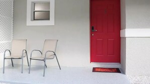 Een rode deur met een klein raam erboven, twee stoelen ervoor en een witte muur.