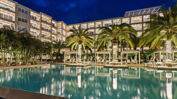 Une piscine entourée de chaises longues et de palmiers, elle-même bordée d’un immeuble d’hôtel à plusieurs étages doté de balcons.