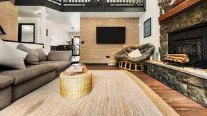 A modern living room with a high ceiling, a stone fireplace, a comfortable sofa, and a flat-screen TV mounted on the wall.
