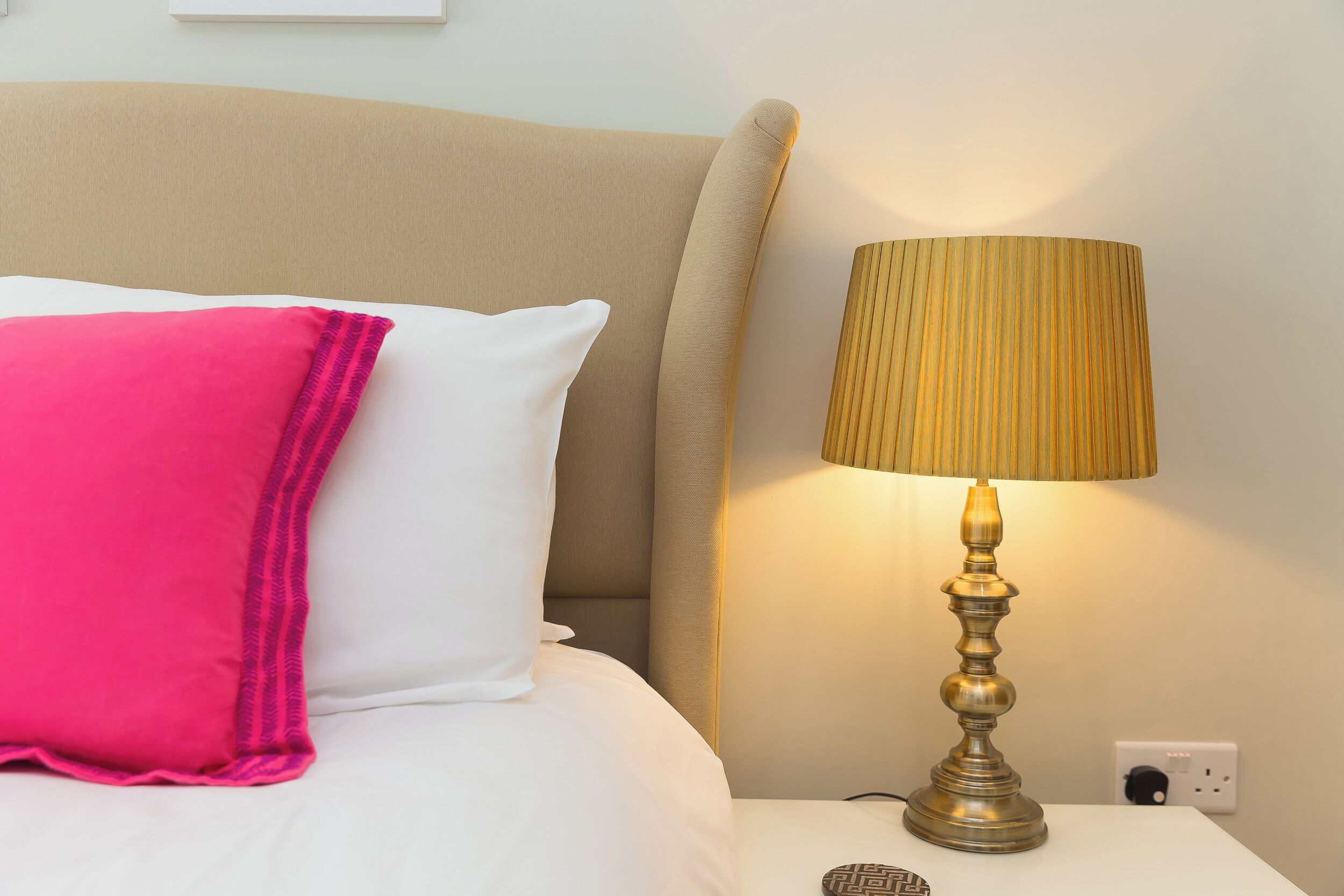 Un lit avec des oreillers blancs et roses, une tête de lit beige et une lampe en laiton sur une table de chevet.