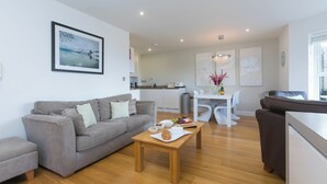 A modern living room with a grey sofa, a wooden coffee table with food, a dining area with a white table and chairs, and a kitchen in the background. - 2 Four Seasons (St Ives)