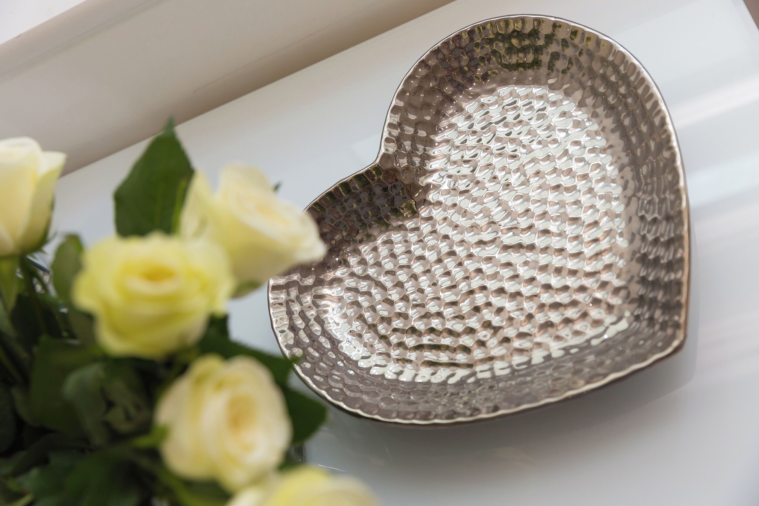 A heart-shaped metallic bowl with a textured surface, placed on a white surface next to a bouquet of yellow roses.