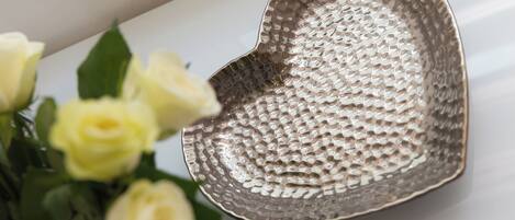 A heart-shaped metallic bowl with a textured surface, placed on a white surface next to a bouquet of yellow roses.