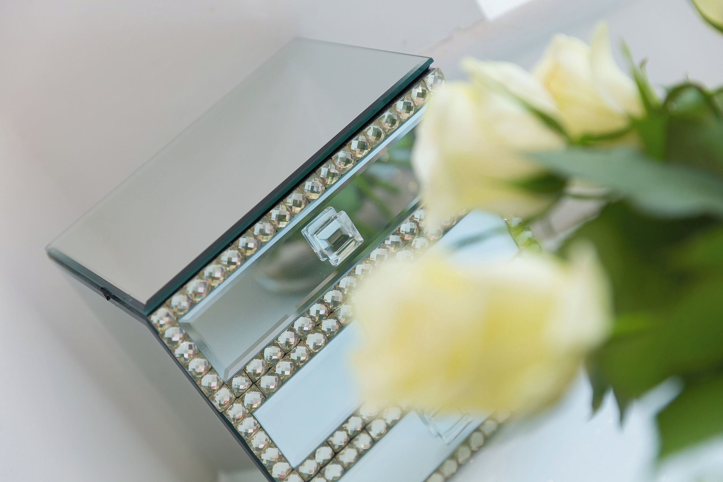 A clear glass mirror with a diamond-studded frame, reflecting a white flower.