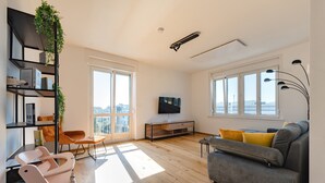 A modern living room with a grey sofa, wooden flooring, a TV on a wooden stand, a bookshelf with plants, and large windows with a city view. - Tamlivingcenterluxus4 Zimmer mit Smart Tvküche (Graz)