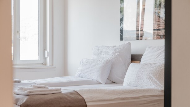 A neatly made bed with white linens and a brown throw blanket, a window with a view of trees, and a painting on the wall. - Tamlivingcenterluxus4 Zimmer mit Smart Tvküche (Graz)