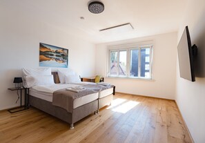 A bedroom with a bed, a bedside table, a lamp, a painting, a window, and a flat-screen TV.