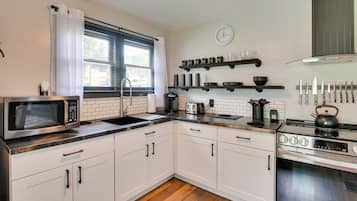 Una cocina moderna con armarios blancos, un microondas de acero inoxidable, una tetera y una estufa. Hay estantes sobre la encimera con vajilla y utensilios.