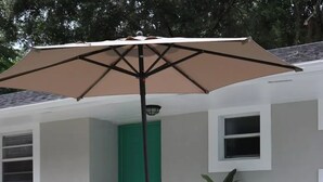 Zona exterior amb seients, dues cadires, un parasol gran, una porta verda i una planta en test.