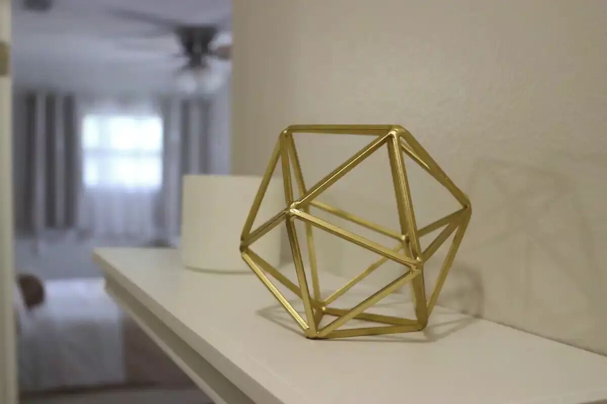 A geometric gold-colored decorative piece on a white shelf in a room with a ceiling fan and a window with curtains.