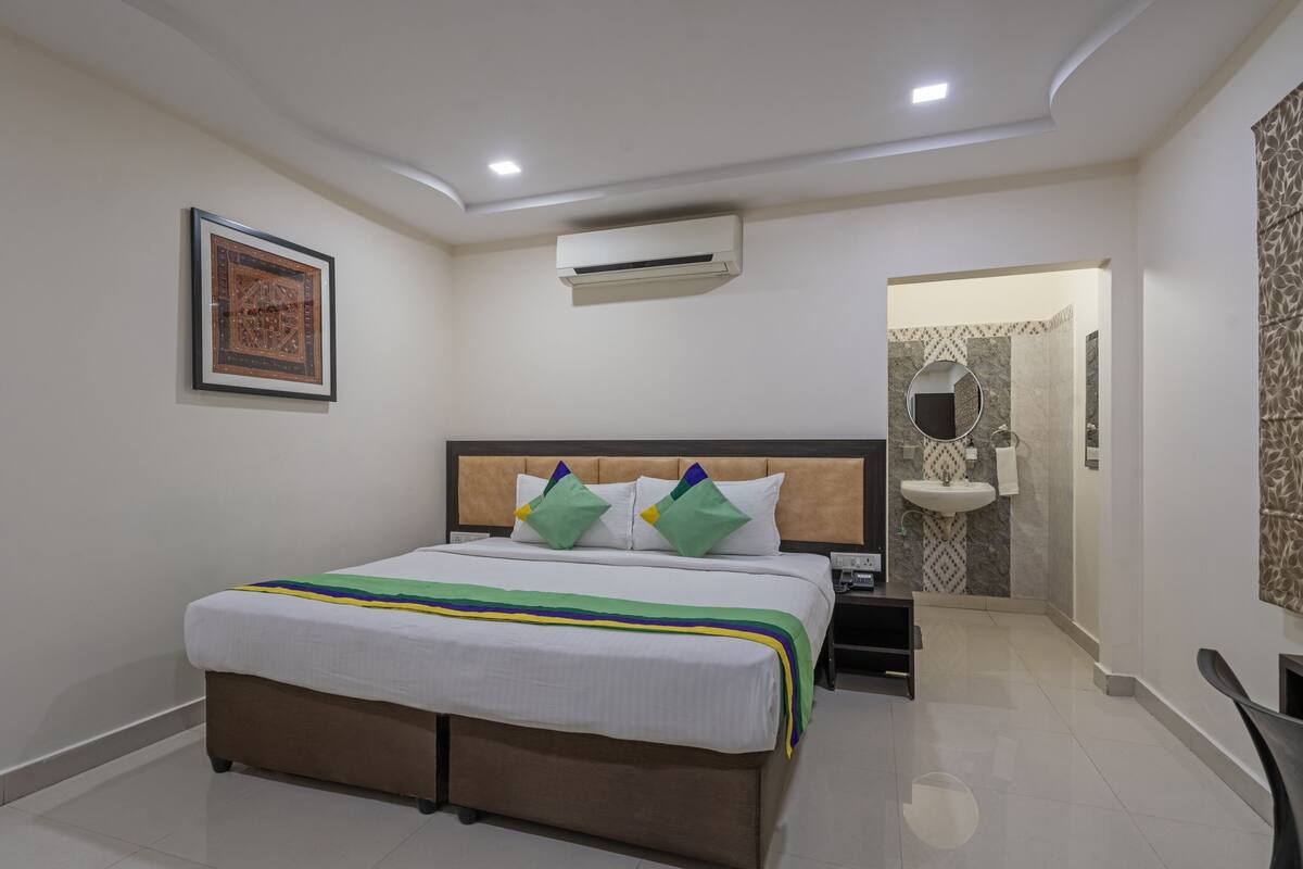 A hotel room with a bed, a nightstand, an air conditioning unit, a bathroom with a sink and mirror, and a framed picture on the wall.