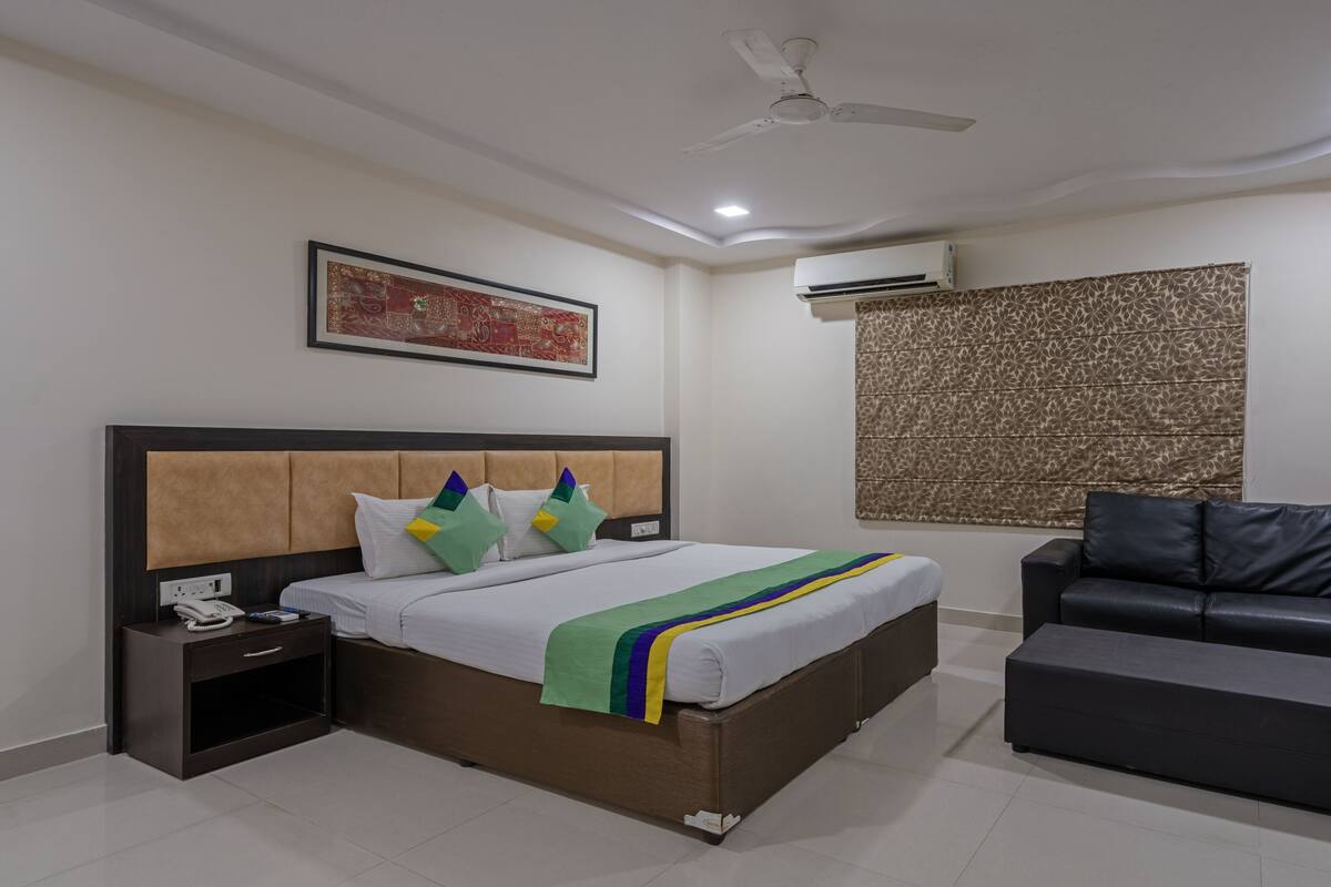 A hotel room with a bed, a bedside table with a phone, a black leather sofa, a patterned wall, and an air conditioning unit.