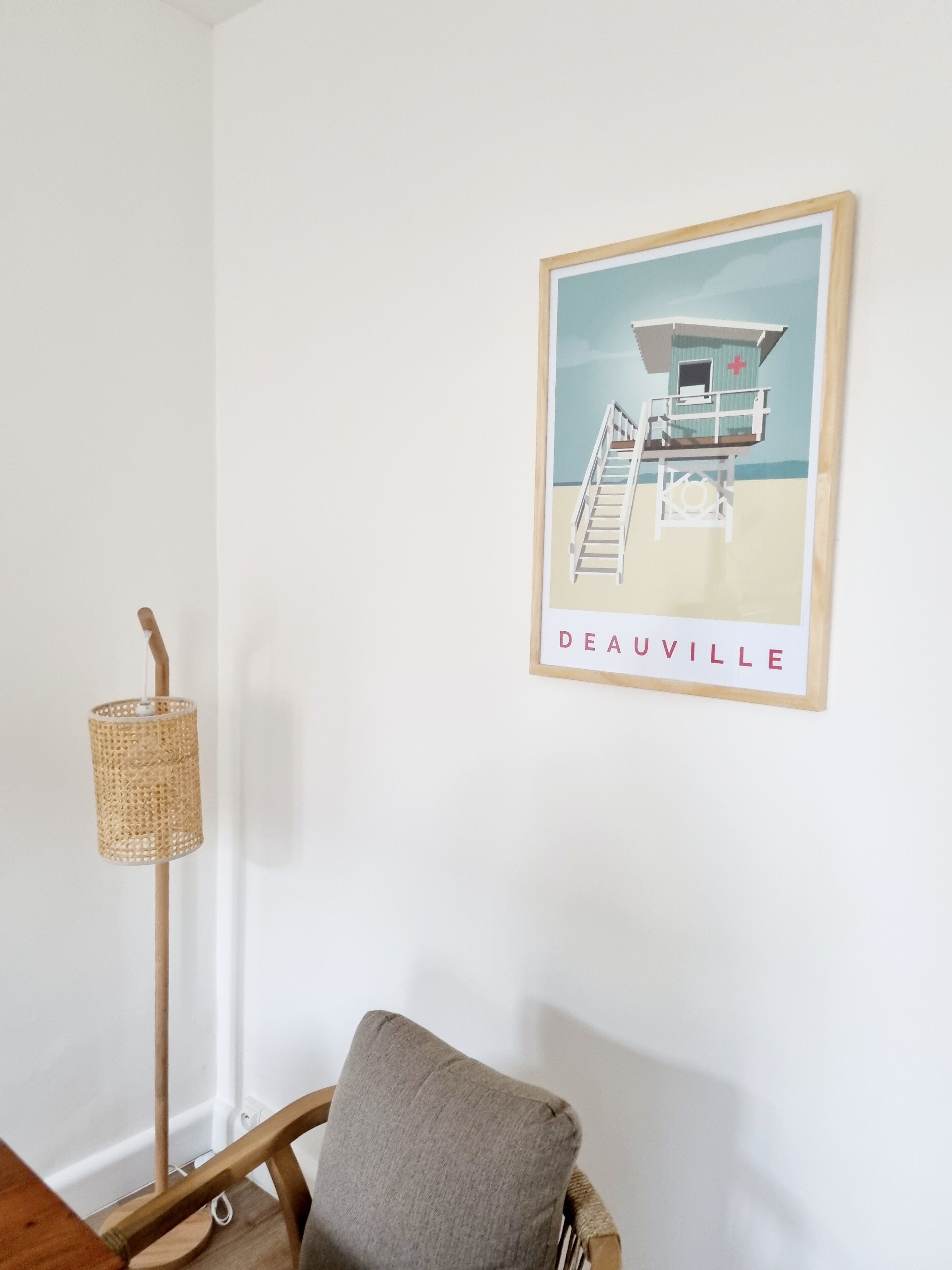 Una habitación con un cartel enmarco de una torre de salvavidas en una playa, una lámpara de pie de mimbre y una silla de madera.