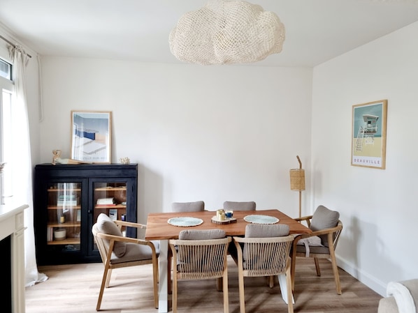 Un coin repas avec une table en bois, six chaises en osier, un buffet noir aux portes vitrées, un luminaire en fibres tressées et deux tableaux encadrés accrochés aux murs.