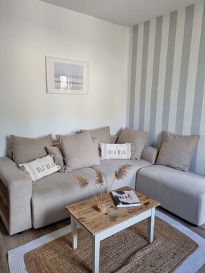 Un salon avec un canapé, deux coussins ornés de l’inscription « BLA BLA », un tableau encadré accroché au mur, une table basse en bois avec un magazine, et un mur à rayures.