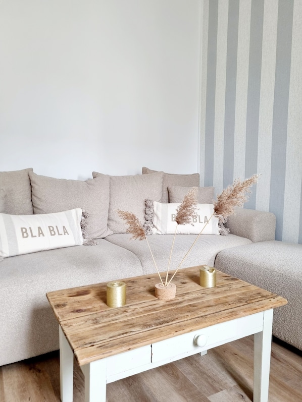 A modern living room with a light grey sofa, two pillows with 'BLA BLA' text, a wooden coffee table with three gold candle holders, and pampas grass in a small vase. - Jolie Maison à 3 min de la Plage et au Pied des Commerces (Blonville-sur-Mer)