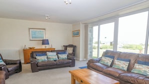 A living room with two brown leather sofas, a wooden coffee table, and a side table with a lamp. There are two framed pictures on the wall and a large window providing a view of the outdoors. - Cim Canol (Pwllheli)