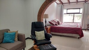 A room with a brown sofa, a massage chair, a bed with a red blanket, and a window with curtains.