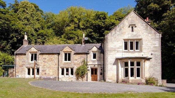 A stone building with a central entrance and two wings, surrounded by a grassy area and trees.