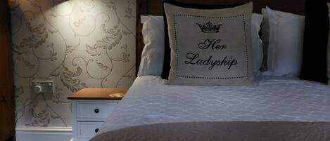 Ein Schlafzimmer mit gemustertem Wandpapier, einem Wanddekor, einer Nachttischlampe, einem Nachttisch und einem Bett mit einem Kopfkissen mit der Aufschrift 'Her Ladyship'.