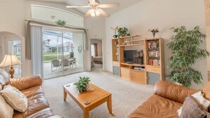 A spacious living room with a ceiling fan, a sliding glass door leading to a patio, a wooden entertainment unit, and a large potted plant.