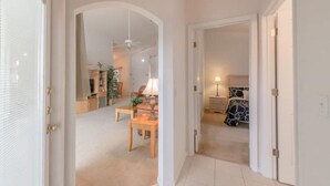 A hallway leading to a bedroom with a bed, a bedside table, and a lamp. There is a door on the left and a window on the right.