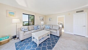 A living room with a beige sofa, a white coffee table, and a patterned rug. There are framed pictures on the wall, a lamp, and a door. - COMPLETELY REMODELED Luxury Riverside 1BR Condo: Panoramic River Views (Marco Island)