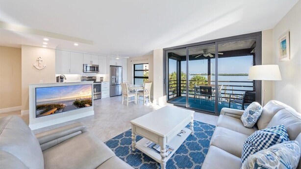 A modern living room with a flat-screen TV, a white coffee table, and a sofa. There is a kitchen area with white cabinets and a dining area with a table and chairs. A large sliding glass door leads to a balcony with a view of the ocean. - COMPLETELY REMODELED Luxury Riverside 1BR Condo: Panoramic River Views (Marco Island)