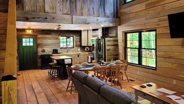 Interior rústico de una cabaña con comedor, cocina y un gran ventanal con vistas a follaje.