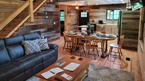 Interior rústico de una cabaña con una escalera de madera, un comedor con mesa y sillas redondas, un sofá de cuero y una zona de cocina.