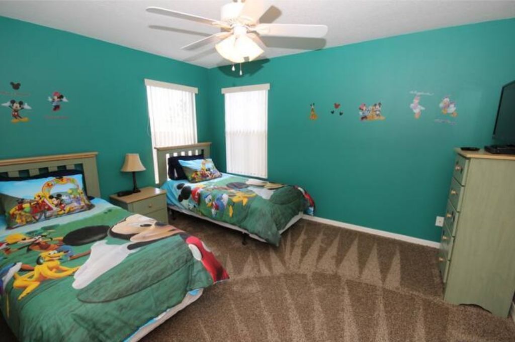 Une chambre avec des murs de couleur turquoise, deux lits avec une literie aux motifs de dessins animés, un ventilateur de plafond et une commode.