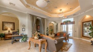A spacious living room with a large sofa, a wooden coffee table, and a desk with a lamp. There are potted plants and a chandelier. - Pool Home in Resort Community (Kissimmee)