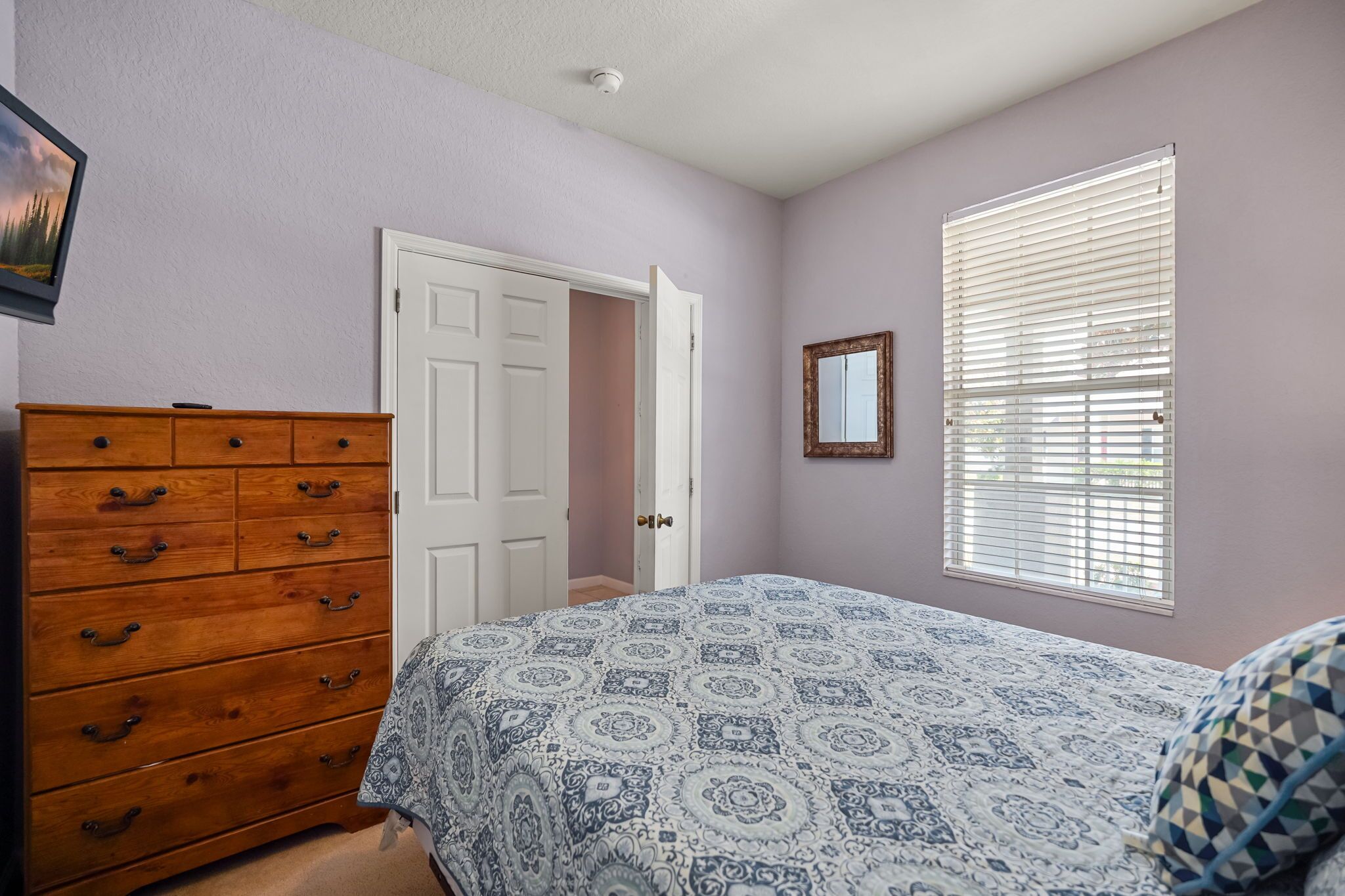 Ein Schlafzimmer mit Bett, Kommode, Tür, Fenster mit Jalousien und einem gerahmten Bild an der Wand.