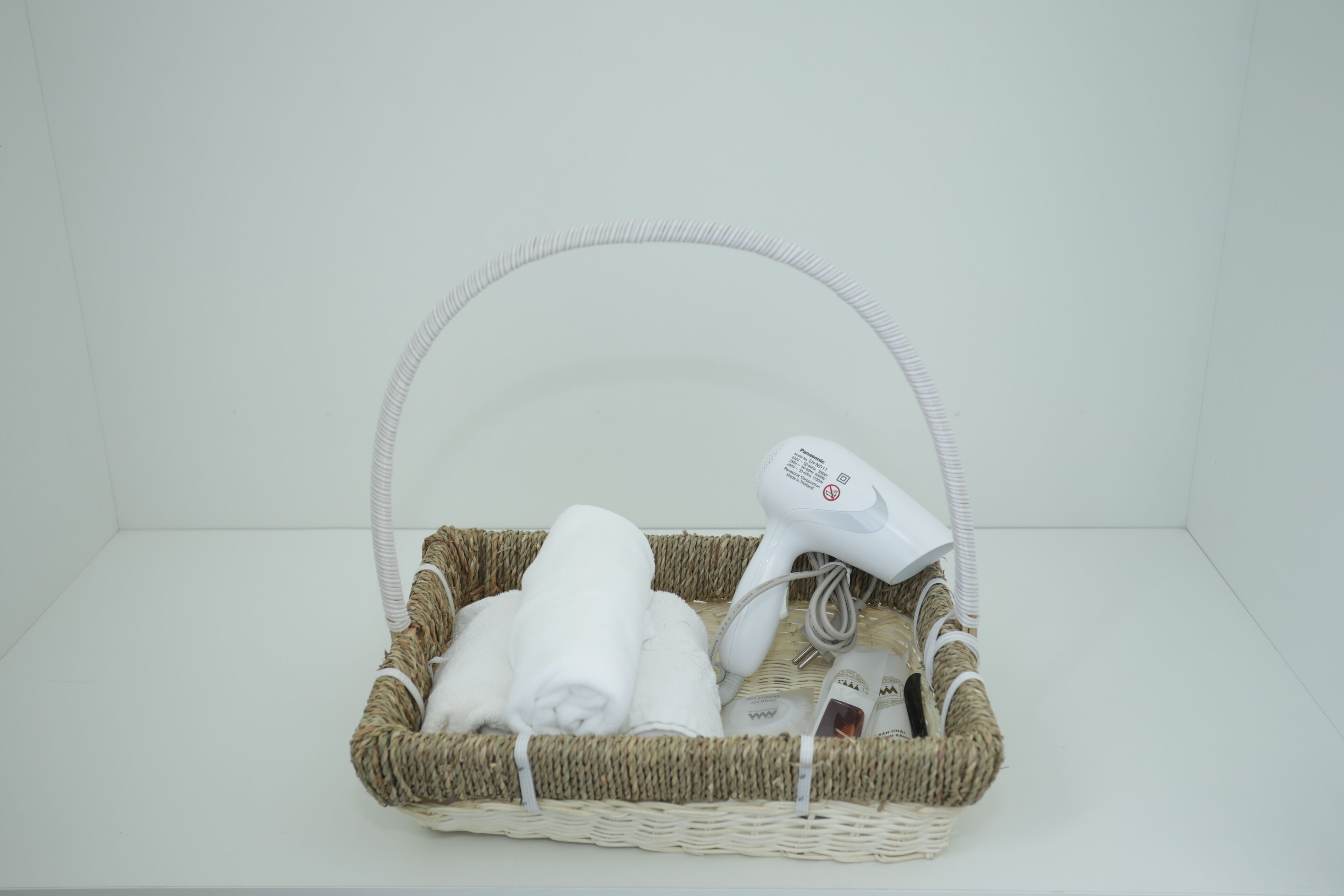 A wicker basket containing a white towel, a hairdryer, and a bottle of lotion.