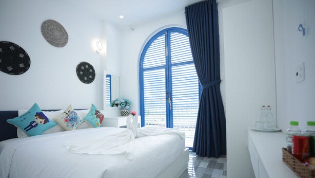 A modern hotel room with a large bed, a small bedside table, a mirror, and a window with blue shutters. - TNJ TASMANIA HOTEL (Da Nang)